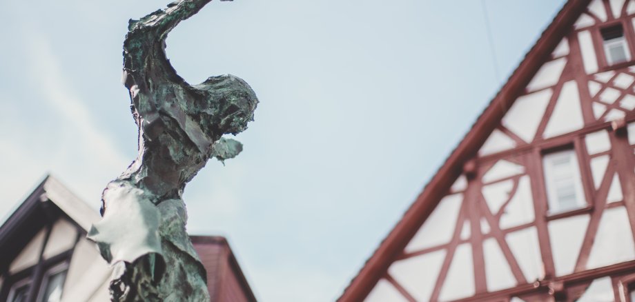 Eine Bronzeskulptur. Im Hintergrund ist ein weiß-rotes Fachwerkgebäude und blauer Himmel zu sehen.