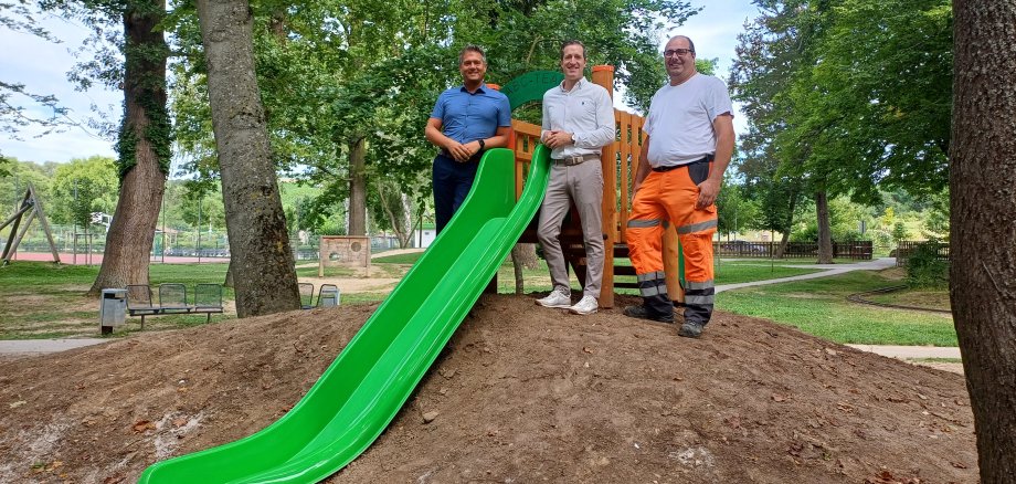 Drei Männer stehen um eine grüne Rutsche für Kleinkinder herum, die auf einem kleinen Hügel aufgebaut ist.