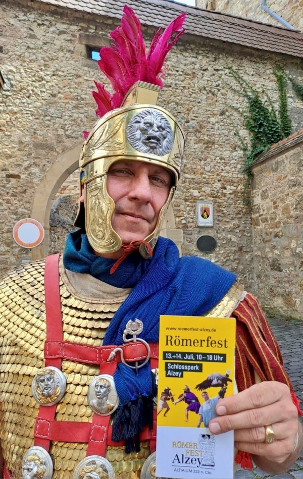 Ein mit einem goldenen Helm, roten Federn und einem silbernen Löwen verzierter römischer Feldherr vor einer alten Sandsteinmauer.