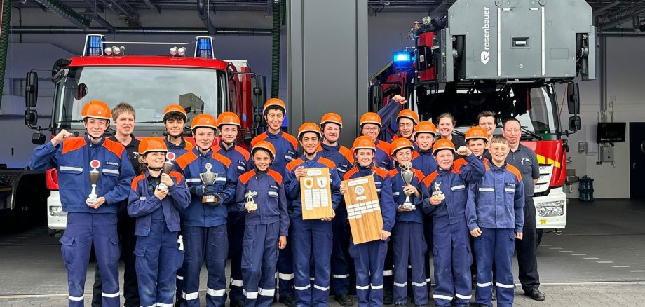 Eine große Gruppe von Kindern und Jugendlichen stehen in ihren blau-orangenen Uniformen vor zwei Einsatzfahrzeugen und zeigen stolz ihre gewonnen Pokale.