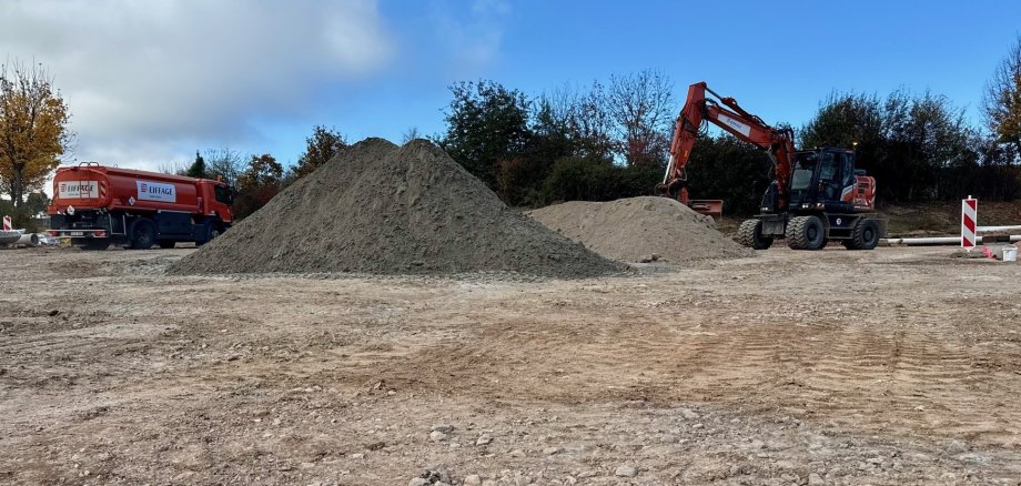 Eine große Schotterfläche mit einem großen Sandberg. Bagger und Lastwagen fahren im Hintergrund.