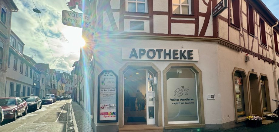 Ein Fachwerkhaus. Über dem Gebäude steht Apotheke. Im Innern stehen Leute an Stehtischen.