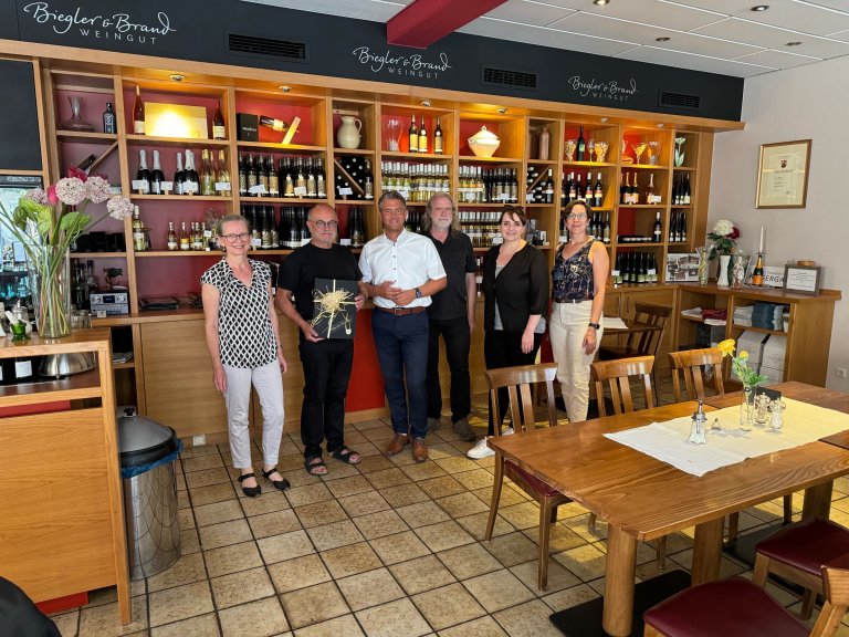 Eine Gruppe von Personen steht vor einem großen Weinregal. Zwei Personen überreichen sich Geschenke.