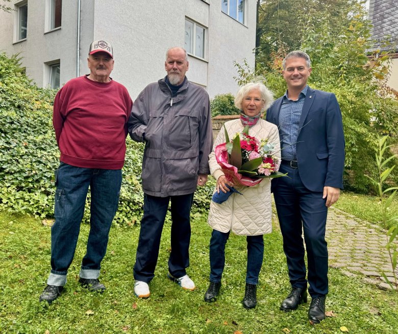 Drei Männer stehen nebeneinander. In ihrer Mitte eine Frau mit einem großen Blumenstrauß.