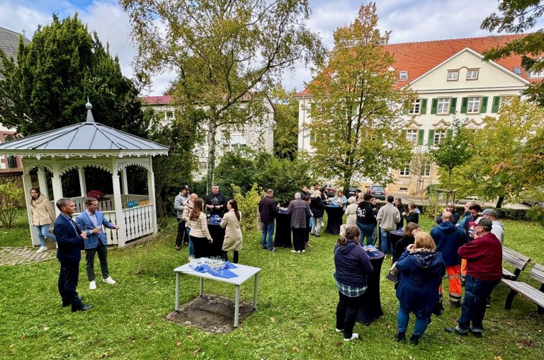 Eine große Grppe von Menschen steht in einem kleinen Park an Stehstischen. Im Hintergrund ist ein kleiner Pavillon zu sehen.