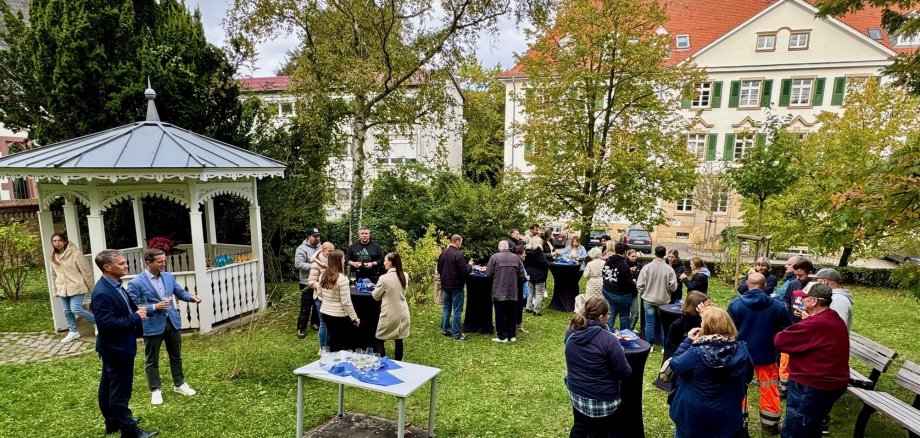 Eine große Grppe von Menschen steht in einem kleinen Park an Stehstischen. Im Hintergrund ist ein kleiner Pavillon zu sehen.